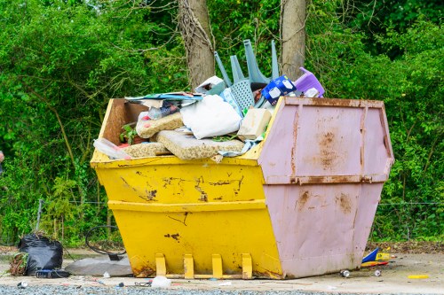 A large, yellow and pink plastic skip filled with various waste items, located outdoors on a gravel surface with green trees and foliage in the background. The skip contains household rubbish, including white plastic bags, cardboard, a blue plastic chair, and other miscellaneous debris. The yellow section of the skip appears weathered with some dirt and scuff marks, while the pink side shows signs of rust and grime. On the ground beside the skip, there are some smaller bags and scattered waste materials. This image depicts typical rubbish collection and disposal setup, relevant to garden clearance services, and highlights the need for professional rubbish removal in Chislehurst or nearby areas. The scene is set in natural daylight, emphasizing the outdoor environment associated with garden and domestic waste management, as might be handled by Garden Clearance Chislehurst.