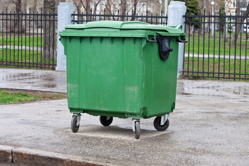 A large green plastic waste bin with a hinged lid and four small wheels is situated on a paved surface outdoors, with a metal fence and a grassy area with trees in the background. The bin's surface appears textured and durable, designed for rubbish collection. Its lid is closed, and the bin is positioned on a damp concrete sidewalk, indicating recent rain. The fence behind features vertical bars, with some concrete posts visible, and the surrounding environment suggests an urban or suburban setting where garden clearance and rubbish removal services, such as those offered by Garden Clearance Chislehurst, are commonly required. The scene is evenly lit, likely by natural daylight, emphasizing the clean and orderly appearance of the bin, which is ready for waste disposal or collection in the local area.