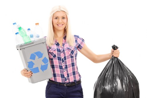A young woman with shoulder-length blonde hair, wearing a pink and blue checked shirt and dark trousers, standing against a plain white background. She is smiling and holding a small gray recycling bin filled with various plastic bottles and containers in her left hand, while her right hand grasps a large black rubbish bag. The recycling bin displays a blue recycling symbol on its side, and the plastic bottles inside are of different colours, including green, clear, and blue, with some caps visible. The black trash bag is tightly gathered at the top. The image appears to be a promotional or educational photograph related to waste disposal, emphasizing recycling and rubbish collection for services possibly offered in Chislehurst, Kent, with a focus on waste management and rubbish removal.