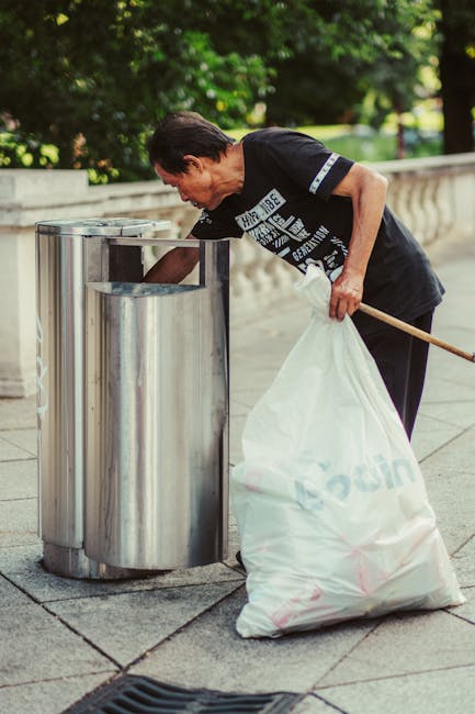 A large pile of mixed waste, including cardboard boxes, plastic bags, paper, and discarded packaging, is overflowing from several outdoor rubbish bins placed on a paved sidewalk in front of a commerci